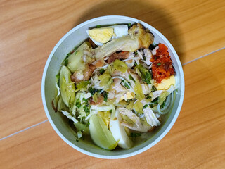 A bowl of Indonesian chicken soto with shredded chicken, sliced boiled egg, vermicelli, sliced cabbage, bean sprouts, celery, chili sauce and lime, mixed with delicious sauce. On a wooden table.