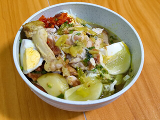 A bowl of Indonesian chicken soto with shredded chicken, sliced boiled egg, vermicelli, sliced cabbage, bean sprouts, celery, chili sauce and lime, mixed with delicious sauce. On a wooden table.