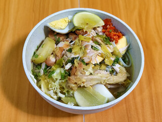 A bowl of Indonesian chicken soto with shredded chicken, sliced boiled egg, vermicelli, sliced cabbage, bean sprouts, celery, chili sauce and lime, mixed with delicious sauce. On a wooden table.