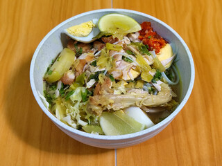 A bowl of Indonesian chicken soto with shredded chicken, sliced boiled egg, vermicelli, sliced cabbage, bean sprouts, celery, chili sauce and lime, mixed with delicious sauce. On a wooden table.