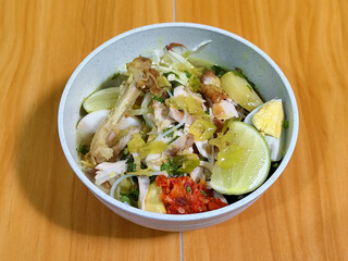 A bowl of Indonesian chicken soto with shredded chicken, sliced boiled egg, vermicelli, sliced cabbage, bean sprouts, celery, chili sauce and lime, mixed with delicious sauce. On a wooden table.