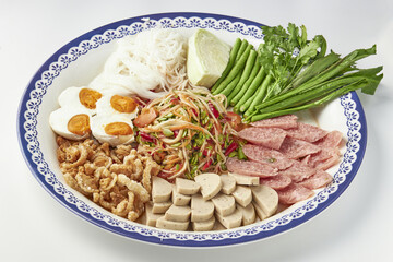 Colorful platter of Thai papaya salad served with variety of side dishes, including boiled eggs, crispy pork rinds, rice noodles, fresh vegetables, and sliced sausages, creating vibrant