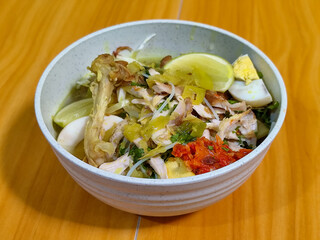 A bowl of Indonesian chicken soto with shredded chicken, sliced boiled egg, vermicelli, sliced cabbage, bean sprouts, celery, chili sauce and lime, mixed with delicious sauce. On a wooden table.