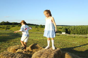 Two children enjoy a joyful afternoon on a farm, climbing and jumping on stacks of hay bales under...