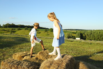 Two children enjoy a playful afternoon in a large green field, climbing on stacked hay bales under...