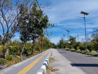 A bright sunny road scene featuring a sidewalk lined with trees and solar street lights under a vivid blue sky with light clouds, capturing the tranquility of a tropical midday