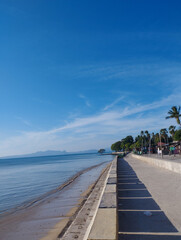 A peaceful beachside walkway stretches along the shore, lined with palm trees and small market stalls. The calm blue sea meets a clear sky, while distant mountains appear faint on the horizon. The str