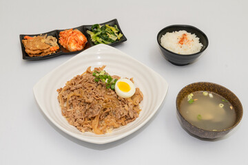 A savory beef rice bowl served as a set meal, featuring tender slices of beef simmered in soy-based sauce atop steamed white rice. The dish is accompanied by a variety of side vegetables and a light