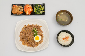 A savory beef rice bowl served as a set meal, featuring tender slices of beef simmered in soy-based sauce atop steamed white rice. The dish is accompanied by a variety of side vegetables and a light