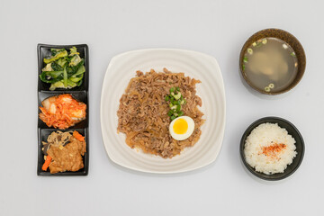 A savory beef rice bowl served as a set meal, featuring tender slices of beef simmered in soy-based sauce atop steamed white rice. The dish is accompanied by a variety of side vegetables and a light