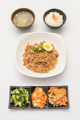 A savory beef rice bowl served as a set meal, featuring tender slices of beef simmered in soy-based sauce atop steamed white rice. The dish is accompanied by a variety of side vegetables and a light