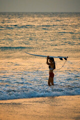 a girl with a surfboard enters the sea at sunset