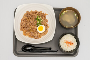 A savory beef rice bowl served as a set meal, featuring tender slices of beef simmered in soy-based sauce atop steamed white rice. The dish is accompanied by a variety of side vegetables and a light