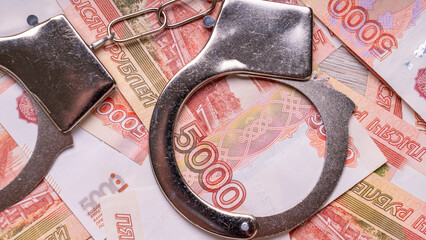 A pair of handcuffs is on top of a pile of money. The handcuffs are silver and the money is red