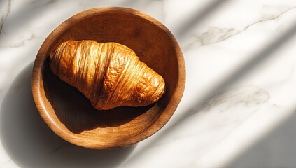 A golden croissant rests in a wooden bowl on a marble surface, bathed in sunlight
