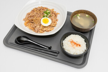 A savory beef rice bowl served as a set meal, featuring tender slices of beef simmered in soy-based sauce atop steamed white rice. The dish is accompanied by a variety of side vegetables and a light