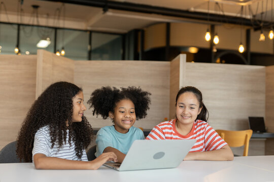 Diverse group of kids three girls happy looking at computer screen together, modern workspace. They are seated at white table while sharing ideas and looking at laptop in classroom school.