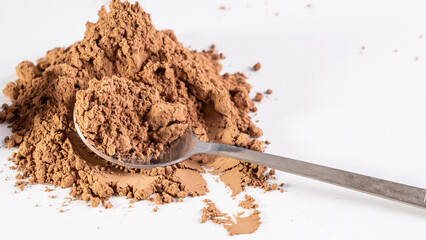 A spoonful of brown powder is piled on a white surface. The spoon is silver and is partially covered in the powder. Concept of abundance and richness, as the spoonful of powder is piled high