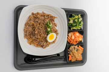 A savory beef rice bowl served as a set meal, featuring tender slices of beef simmered in soy-based sauce atop steamed white rice. The dish is accompanied by a variety of side vegetables and a light