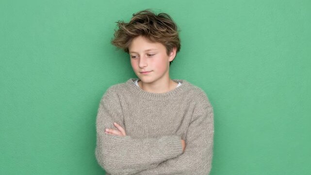 Caucasian boy with messy hair in gray sweater on green background. Child model posing with crossed arms. Studio portrait for youth fashion, confidence concept - Powered by Adobe