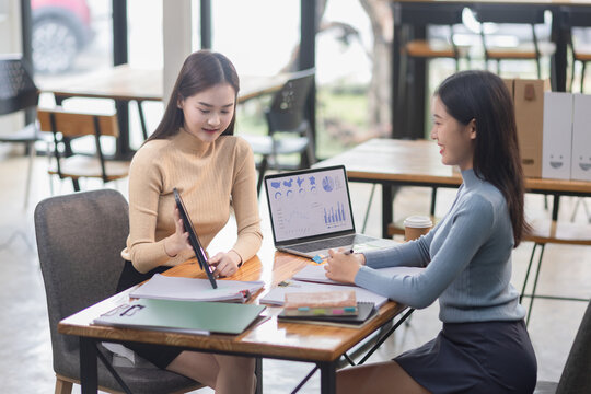 Two asian Businesswoman using tablet and laptop reviewing digital financial graphs and reports working in office desk.