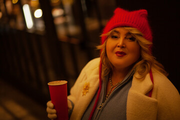 Plus size woman walks around the city during the Christmas of the week with a hot drink in a glass against the background of flickering light bulbs