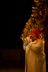 Plus size woman walks around the city during the Christmas of the week with a hot drink in a glass against the background of flickering light bulbs