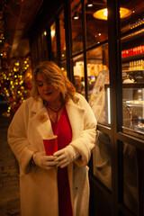 Plus size woman walks around the city during the Christmas of the week with a hot drink in a glass against the background of flickering light bulbs