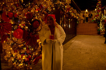 Plus size woman walks around the city during the Christmas of the week with a hot drink in a glass against the background of flickering light bulbs