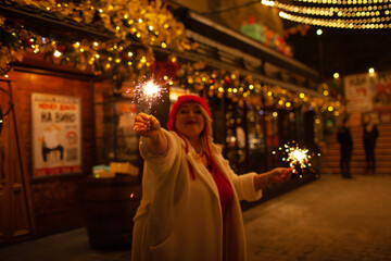 Plus size woman walks around the city during the Christmas of the week with a hot drink in a glass against the background of flickering light bulbs