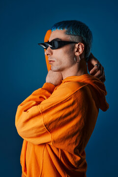 Side view of confident male model in prism glasses and bright orange hoodie standing on blue background in studio and looking away