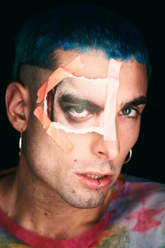 Handsome male model with blue hair and cutout printed photo paper eye stuck on face looking at camera on black background in studio
