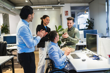 Obraz premium Group of happy young entrepreneurs clapping their colleague in a wheelchair, celebrating a success in a modern coworking office
