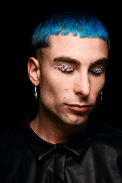 Extraordinary delighted male model with blue hair and eye stickers on face looking at camera on black background in studio