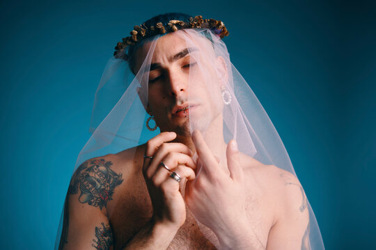 Male model with blue hair and naked torso standing in white transparent veil on blue background in studio and looking at camera
