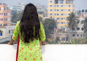 Bengali woman in embroidered green salwar kameez, long black hair flowing, stands alone on a rooftop. Back view captures her looking out, a moment of solitude. Authentic South Asian culture.