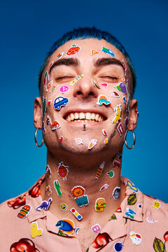 Naklejki Content male model with various colorful stickers on face and with blue hair smiling with closed eyes in studio