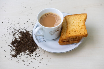 Hot milk tea, also known as dudh cha, with delightful bubbles in a teacup. Crispy rusk biscuits alongside. Fresh and dried tea leaves artfully arranged on a wooden surface.