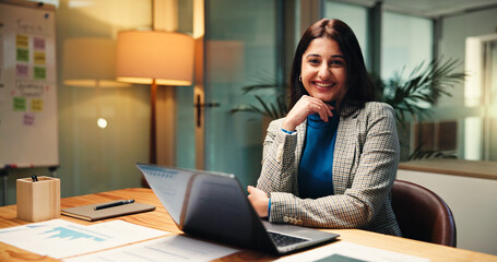 Laptop, portrait and smile of business woman in office at night for social media analytics. Computer, development and statistics with happy Indian employee at desk in workplace for evening deadline