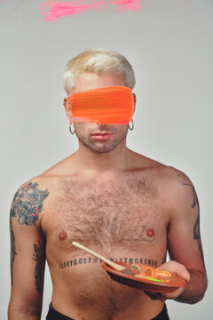 Young male model with tattooed hairy torso and stroke of paint on eyes standing behind glass against gray background