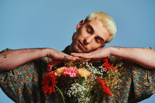Trendy young guy in ornamental shirt touching bunch of natural flowers and looking at camera against blue background
