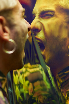 Enraged young male with earring yelling in dissatisfaction at reflection on dichroic film