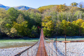 An iron suspension bridge on steel cables across a mountain river leading to the forest on a sunny autumn day