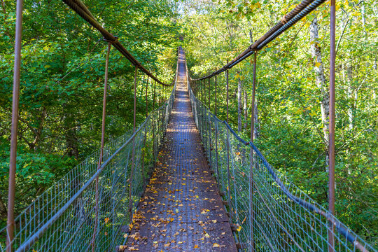 An iron suspension bridge on steel cables across a mountain river leading to the forest on a sunny autumn day