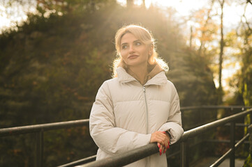 Young woman in a white puffer jacket walking through a beautiful green natural park on a sunny day. Outdoor lifestyle, peaceful moment, nature and fashion concept.