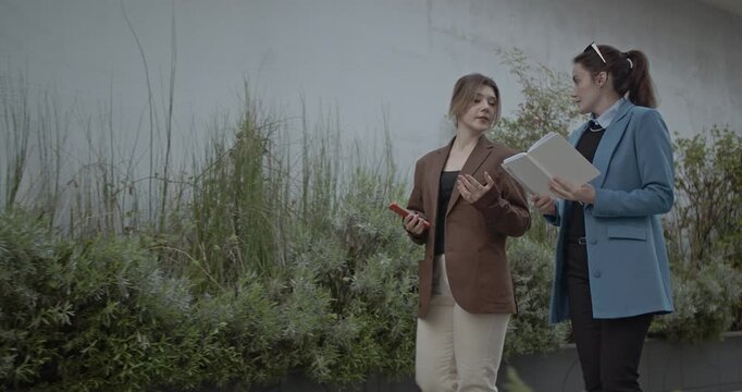 Two young businesswomen walk and talk, discussing something in a notebook.