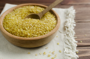 Raw bulgur wheat grains in a wooden bowl with a spoon on a napkin on a brown table. Healthy eating concept. Vegan and vegetarian food. Diet. Rustic style. Horizontal orientation. Copy space