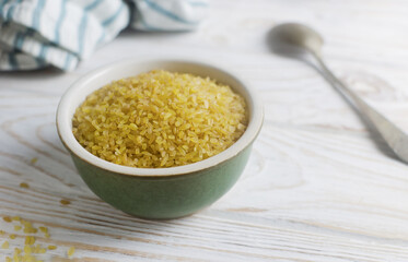Raw bulgur wheat grains in a ceramic green bowl with a spoon and napkin on a white wooden table. Healthy eating concept. Vegan and vegetarian food. Rustic style. Vertical orientation. Copy space