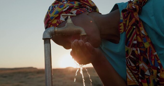 Backlit cropped close-up of a Black African woman in traditional clothing drinking water from a faucet at sunset