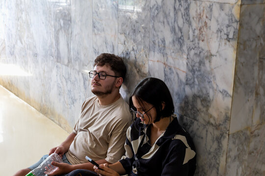 Exhausted young people sitting near textured marble wall. Girl focused on smartphone. Guy holding empty bottle from under water. Tired tourists, man and woman, sitting on floor and resting